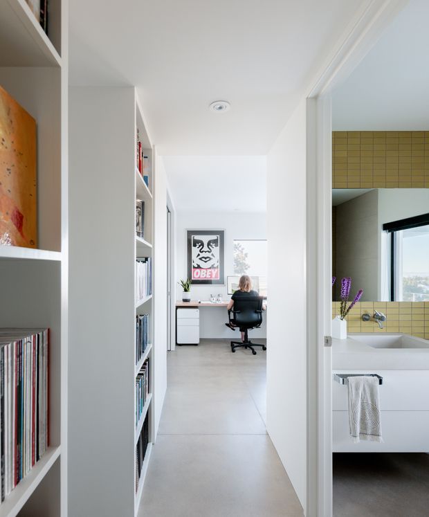 Bright, minimalist interior view down a hallway lined with built-in white shelving and books. A compact home office with a desk and chair is visible at the end, while an open bathroom with a white vanity, basin, black-framed window, and mustard-yellow grid tile splashback is visible on the right.