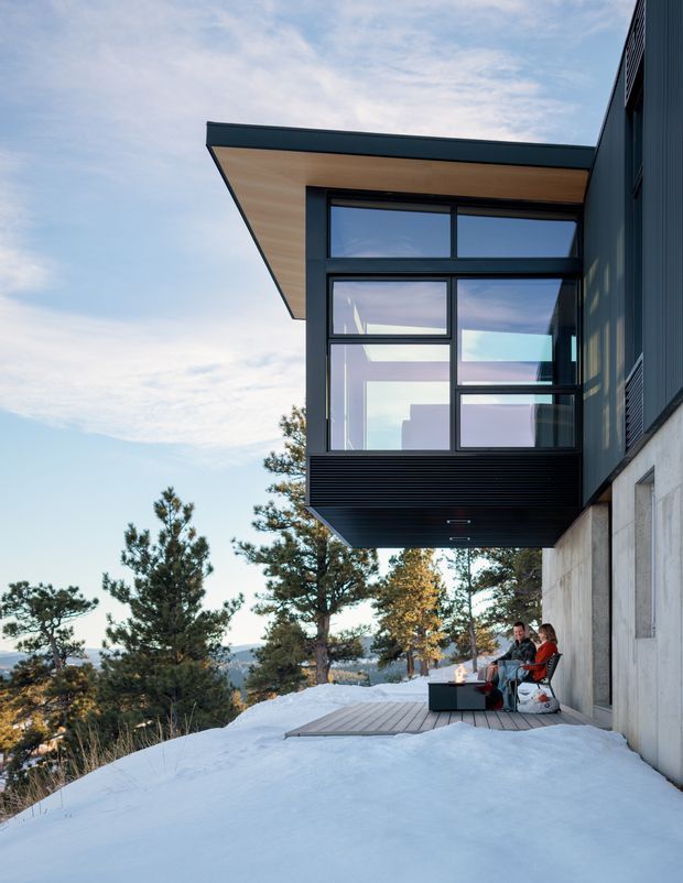 Exterior architectural photograph of a contemporary, cantilevered building set on a snowy, wooded sloped site. The dark-clad structure has a projecting flat roof, extensive black-framed glazing, exposed concrete lower walls, and a timber deck with outdoor seating beneath the overhang. Pine trees, snow-covered terrain, and distant mountainous scenery surround the building.