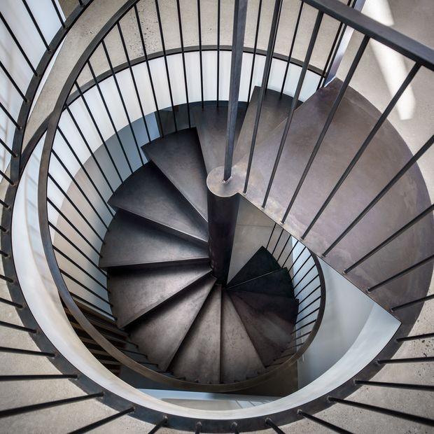 Top-down view of a sculptural spiral staircase with dark metal or concrete treads, a central cylindrical column, and slender black metal balustrades. The staircase curves through a bright, modern circular stairwell with pale walls and a textured concrete ceiling.