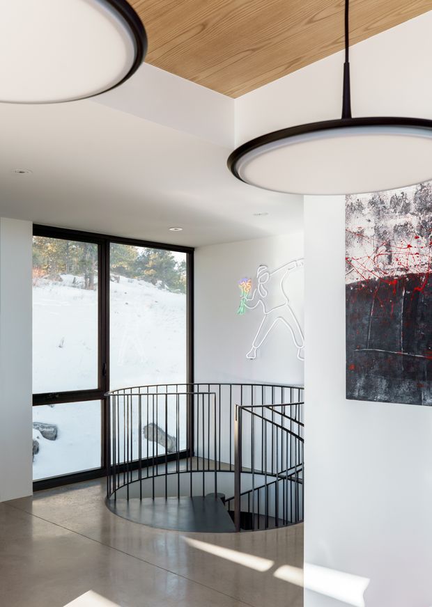A bright contemporary interior landing overlooking a circular stairwell. The space features white walls, polished concrete flooring, large black-framed windows with snowy outdoor views, slim black metal balustrades, a timber-lined ceiling section, modern circular pendant lights, and abstract wall artwork including a neon-style outline figure.