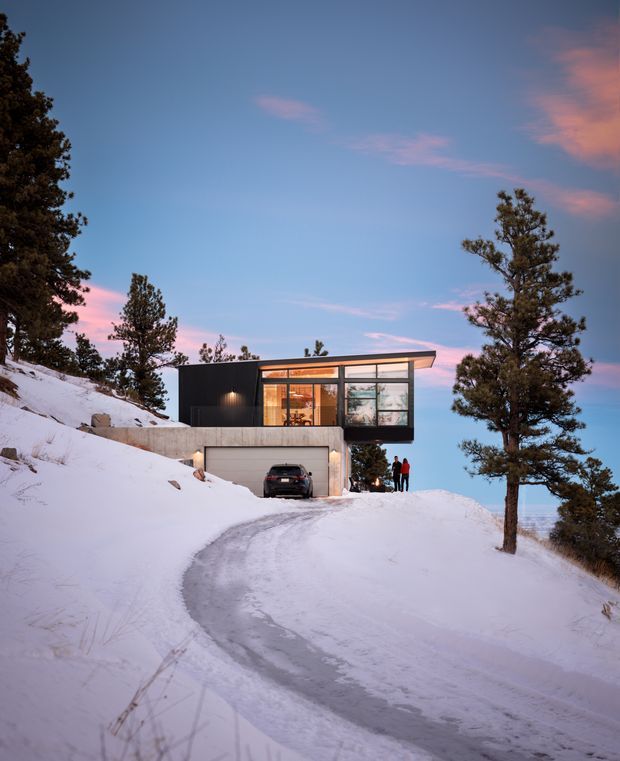 Exterior view of a compact contemporary house set into a steep, snow-covered wooded hillside. The home has a dark, rectilinear upper volume with extensive floor-to-ceiling glazing, a projecting flat roof, and a concrete garage level beneath. A curved driveway leads up to the integrated garage, with pine trees and snowy terrain surrounding the property at dusk.