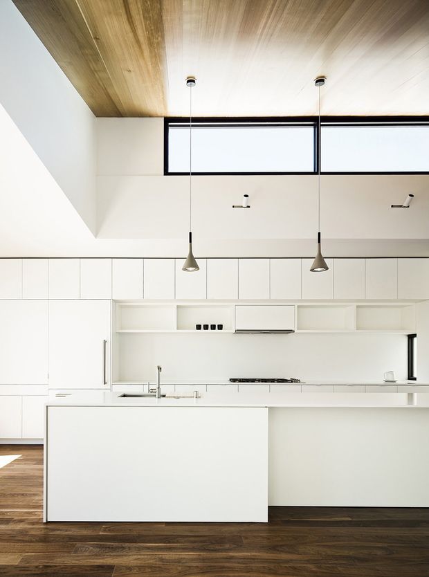Bright minimalist kitchen with extensive white built-in cabinetry, a large white island, integrated cooktop and sink, and open shelving. The room has dark timber flooring, a timber-lined ceiling, black-framed clerestory windows, and two pendant lights. The high ceiling and clerestory glazing create an airy, modern interior.