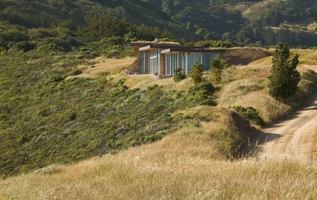 A small, low-profile modern dwelling embedded in a dry, vegetated hillside. The building has extensive floor-to-ceiling glazing, simple rectilinear volumes, a flat or very low-pitched roof, and warm timber-toned exterior elements with dark masonry visible at the rear. A narrow unpaved driveway approaches from the right, while rolling hills and native scrub surround the property.