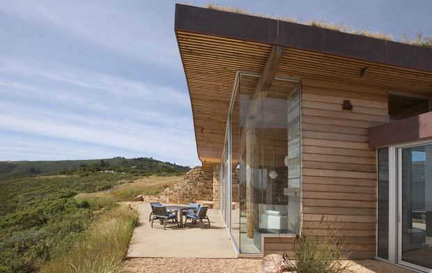 Low-profile contemporary single-storey house set into a sloping rural landscape, with broad floor-to-ceiling glazing, warm horizontal timber cladding, a deep timber-lined roof overhang, exposed structural beams, and a paved outdoor terrace. The flat roof has a planted or grass-covered edge, while stonework integrates the building into the site. Rolling hills and native vegetation surround the home.