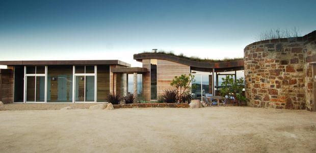 Low-slung contemporary single-storey residence arranged around a broad sandy courtyard. The building combines horizontal timber-clad volumes, extensive floor-to-ceiling glazing with dark frames, gently curved roof forms with planted roof edges, and a prominent rough natural-stone wall on the right. Sparse drought-tolerant landscaping and an outdoor seating area reinforce the restrained modern setting.