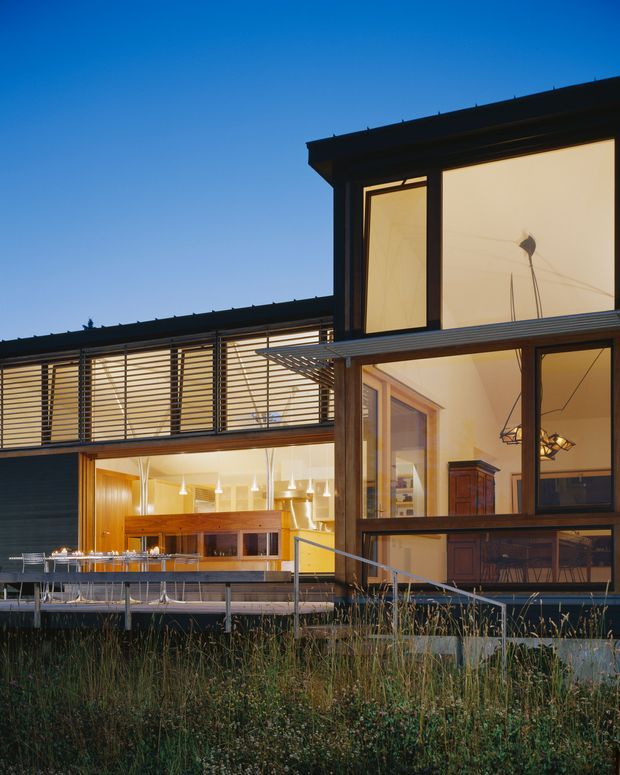 Exterior dusk view of a contemporary multi-level residence with dark-framed floor-to-ceiling glazing, strong horizontal lines, timber-lined interior spaces, and an elevated outdoor dining terrace. Warm interior lighting reveals a kitchen and dining/living areas through the glass, while landscaped grasses occupy the foreground.