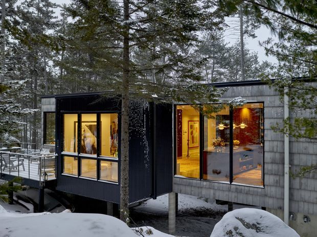 Exterior view of a contemporary, elevated house in a snowy forest setting. The compact structure combines dark ribbed cladding with grey shingle-like wall panels and extensive floor-to-ceiling glazing, revealing warmly lit interior spaces. A deck and railings extend to the left, while the building is supported above the snow-covered ground among mature trees.