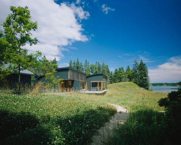 Exterior view of a low, modern timber-clad house set in a natural grassy landscape beside a body of water. The building has dark horizontal cladding, large timber-framed windows, simple low-pitched roof forms, and a path leading through meadow vegetation toward the home. Forest and shoreline are visible in the background under a bright blue sky.