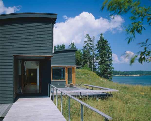 Exterior view of a contemporary dark-clad building beside a calm body of water. The low, flat-roofed structure has horizontal cladding, timber-framed glazing, a timber deck and a simple metal balustrade extending toward the waterfront. Native trees and grasses surround the building under a bright blue sky.