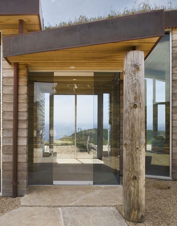 Exterior view of a contemporary timber-clad building entrance, featuring a glazed entry vestibule, broad projecting canopy with a planted roof, exposed timber soffit, and a large rough-hewn timber post. The surrounding gravel landscape and distant coastal horizon are visible through the glazing.