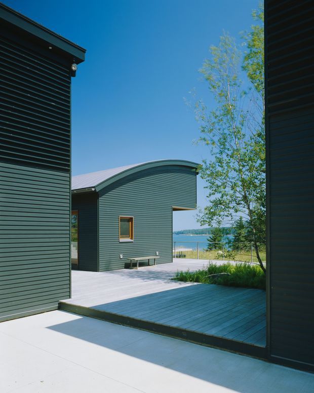 Exterior architectural view through a narrow passage between dark green horizontally clad building volumes. A curved-roof pavilion sits beyond a broad timber deck and pale concrete paving, with a leafy tree and a visible body of water in the background under a clear blue sky.