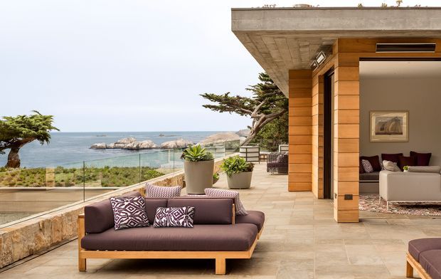 Contemporary coastal terrace overlooking the ocean, with a low-profile outdoor sofa, patterned cushions, potted plants, glass balustrade, and natural stone paving. A timber-clad living room pavilion with broad roof overhang opens directly onto the terrace, while rocky shoreline and landscaped coastal terrain are visible beyond.