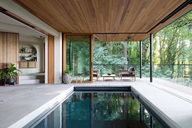 A refined indoor pool area with a narrow rectangular swimming pool, surrounded by pale concrete or stone flooring and extensive floor-to-ceiling glazing. The space opens directly to a forested setting and includes a timber-lined ceiling, dark-framed glass walls, and a small minimalist lounge seating area. An adjacent room with built-in shelving and timber detailing is visible to the left.