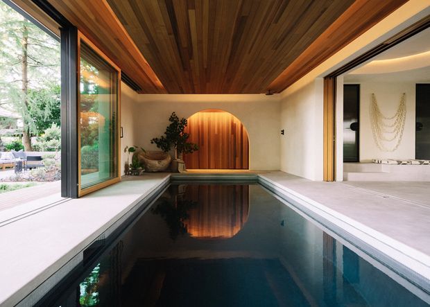 A refined indoor pool room with a narrow lap pool extending toward a curved timber feature doorway. The space has warm wood-lined ceilings and feature joinery, pale plaster walls and flooring, large black-framed glazed openings connecting to a landscaped outdoor area, and an adjacent minimalist room visible through open glazing. The composition emphasizes a calm, resort-like indoor-outdoor connection.