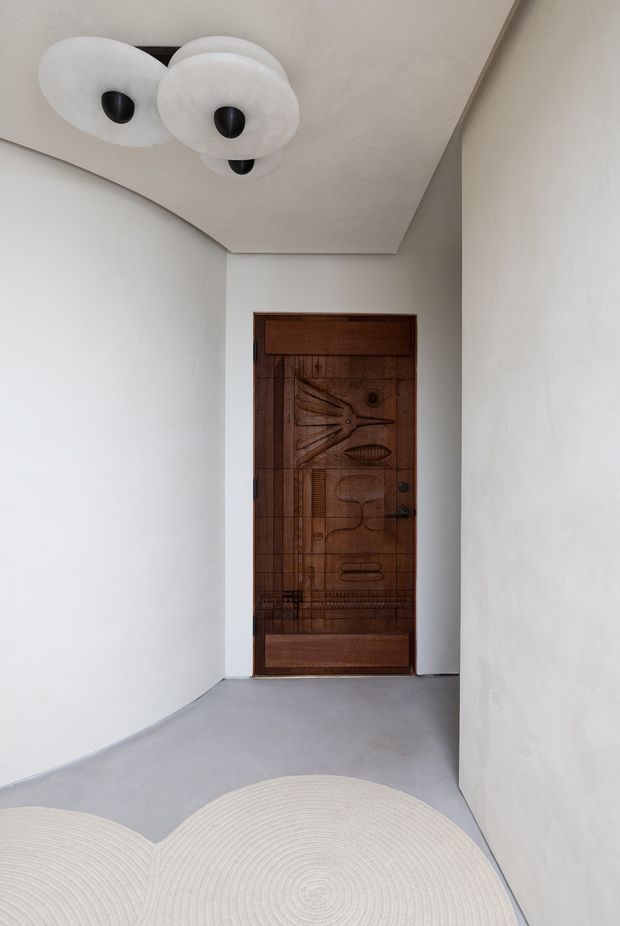 A minimalist residential entryway with smooth white plaster walls, a pale grey floor, and a highly distinctive dark timber front door featuring carved geometric and organic relief motifs. A sculptural ceiling light with layered white discs and black central fittings adds a decorative focal point, while a curved cream rug softens the foreground.