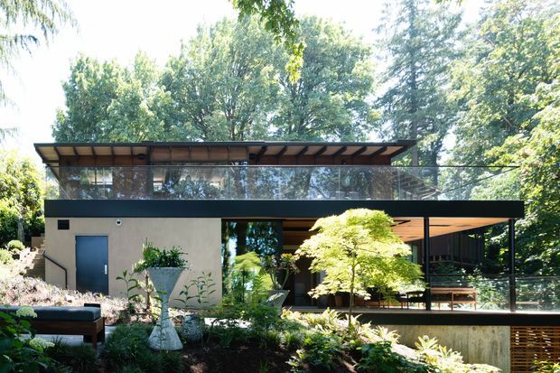A contemporary two-storey residential house set within mature landscaped greenery. The building features extensive floor-to-ceiling glazing, dark horizontal framing, a broad low-pitched roof canopy, warm timber soffits, and an open covered terrace or carport-like space. Layered garden planting and a sculptural outdoor planter sit in the foreground.
