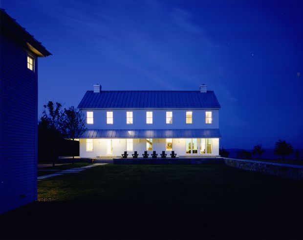 Twilight exterior view of a large two-storey white house with a simple rectangular form, dark standing-seam pitched roof, regularly spaced illuminated windows, and a bright glazed ground-floor living area opening toward a lawn and terrace. A darker adjacent building is partially visible at the left edge, with a low stone wall extending across the right foreground.