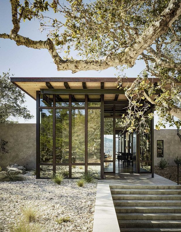 Exterior view of a low-slung modern house set within a dry, landscaped garden. The entrance is framed by extensive floor-to-ceiling glazing in dark metal frames, with a projecting flat roof, exposed timber soffit and dark structural beams. Broad concrete steps lead through a glazed entry into the interior, while mature trees and a pebble-covered landscape surround the building.