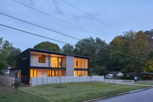 A two-storey contemporary flat-roof residence viewed from across a suburban street at dusk. The dark-clad rectangular building has extensive warm-lit glazing, a projecting upper-level balcony, pale vertical wall panels, and a low concrete boundary wall. A broad lawn, driveway, neighbouring homes, mature trees, and overhead utility lines surround the property.