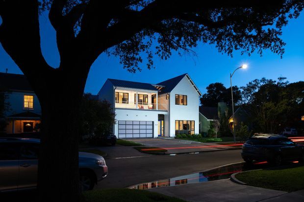 Twilight street view of a contemporary multi-level residential house with pale horizontal cladding, a steep gabled roof, large upper-level windows, a glazed balcony or porch, and an integrated double garage. Mature trees frame the foreground, with neighbouring homes, street lighting, parked cars, and wet pavement visible around the property.