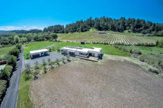 Aerial view of a contemporary single-storey rural property surrounded by expansive cultivated fields, vineyard-like planted rows, lawns, mature trees, and a private driveway. The main residence has a low horizontal form with a flat roof, dark and light exterior finishes, and an adjacent detached outbuilding or garage.
