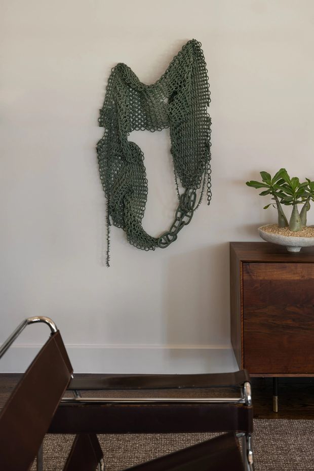 A refined interior vignette featuring a dark wood mid-century-style sideboard, a sculptural green metal chain wall artwork, a potted plant, and a dark leather-and-chrome lounge chair in the foreground. The space has warm neutral walls, dark timber flooring, and a textured natural-fibre rug.