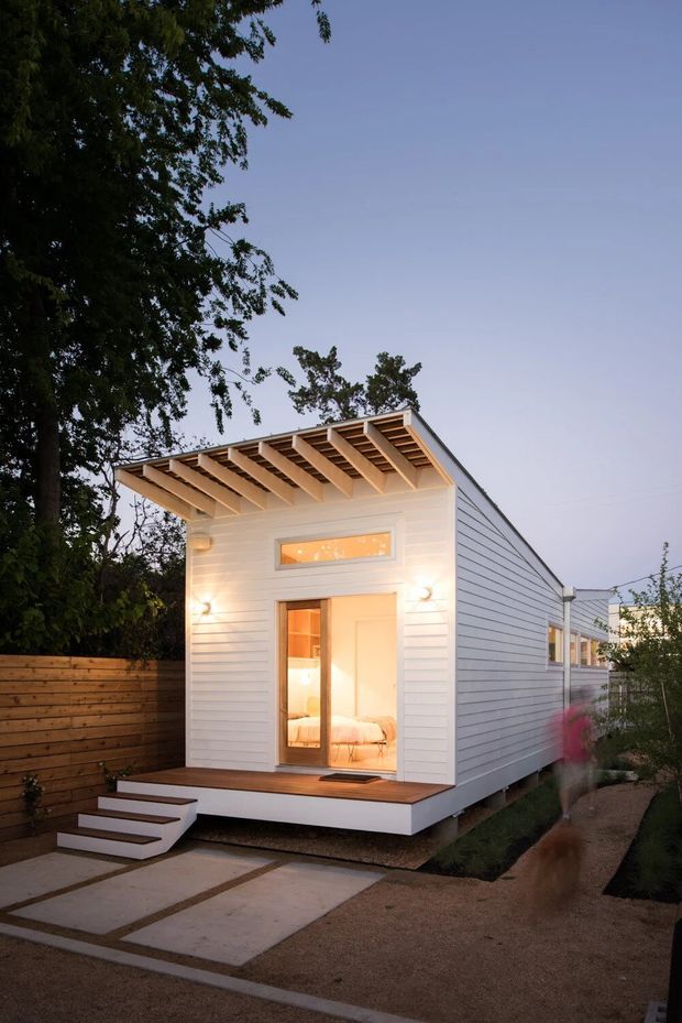 Exterior view of a compact contemporary backyard dwelling or studio with white horizontal cladding, a projecting timber pergola-like roof structure, glazed entry, and a small raised timber deck with steps. The building sits along a landscaped walkway beside a timber boundary fence and mature trees, with warm interior lighting visible through the door and clerestory window.