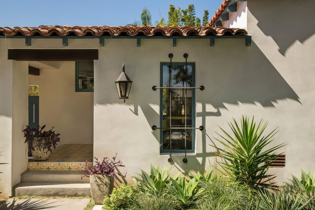 Sunlit exterior facade of a stucco residence with a terracotta tiled roof, deep roof eaves, dark timber or metal window and door frames, decorative wrought-iron window grilles, and a traditional wall-mounted lantern. A recessed entry porch with patterned tile risers opens into the home, while drought-tolerant landscaping with spiky ornamental plants fills the foreground.