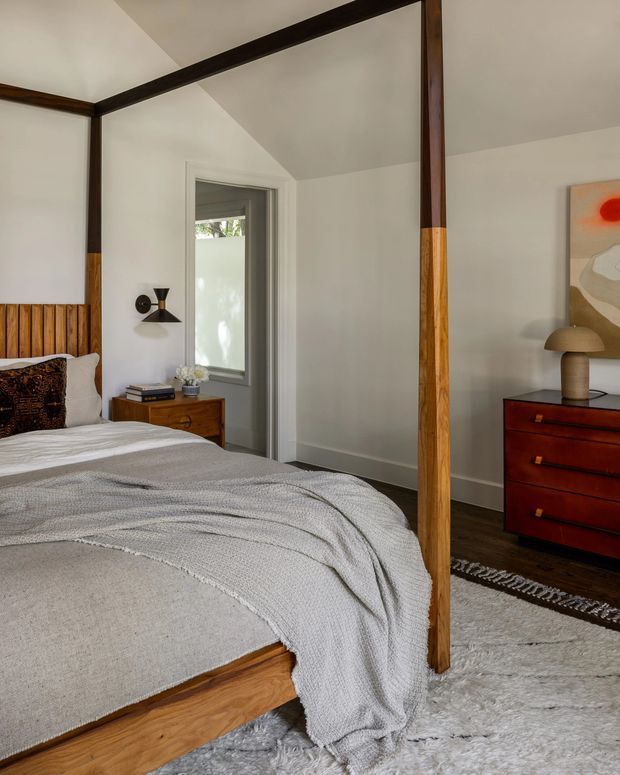 A warm, contemporary bedroom featuring a timber four-poster bed with dark-stained upper framing and natural wood posts, layered white and grey bedding, a timber slatted headboard, and dark timber flooring. A large wall mirror reflects the bed and doorway. The room includes a mid-century-style red timber dresser, bedside table, sculptural wall sconce, table lamp, artwork, and a pale textured rug.