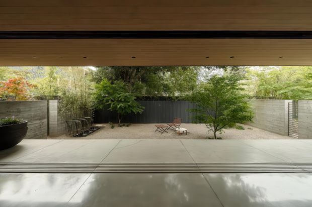 A calm, sheltered courtyard viewed from beneath a deep timber-lined roof or covered indoor-outdoor opening. Large smooth concrete or stone paving extends toward a gravel garden with mature planting, concrete boundary walls, a dark vertical metal privacy fence, and simple outdoor chairs and a small table. The composition emphasises a strong connection between interior and landscaped exterior, with a low horizontal roofline and restrained contemporary detailing.