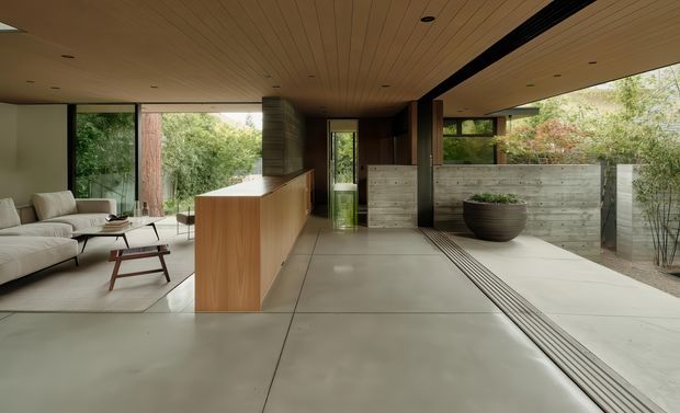 A refined open-plan interior with a minimalist kitchen island, lounge area, and extensive glazing opening toward a landscaped courtyard. The room features a warm timber-lined ceiling, timber kitchen cabinetry, smooth large-format pale concrete or stone flooring, dark structural framing, and a restrained neutral palette. A concrete privacy wall and planted courtyard are visible beyond the glazing.