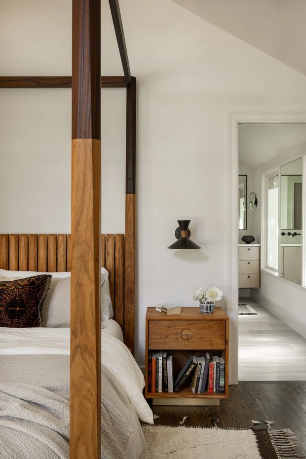 A warm, refined bedroom with a substantial timber four-poster bed, white bedding, patterned accent cushion, and a timber bedside cabinet filled with books. The room has white walls, dark timber flooring, a textured neutral rug, and a sculptural black wall sconce. A bathroom or dressing area is visible through the doorway and mirror at right.