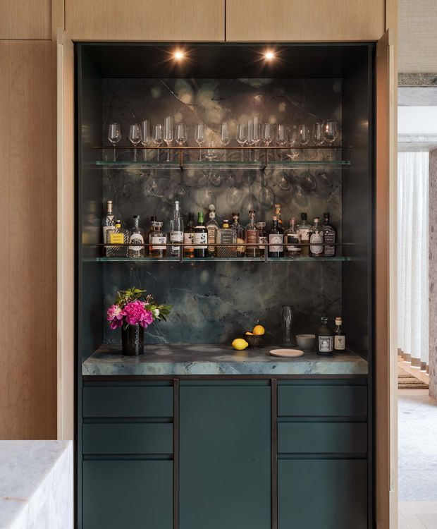 A compact built-in home bar recessed into full-height timber cabinetry. The bar features dark green lower cabinetry, a richly veined dark stone benchtop and matching stone backsplash, glass shelves displaying wine glasses and liquor bottles, and integrated overhead spotlights. A doorway to a bright adjacent room is visible on the right.