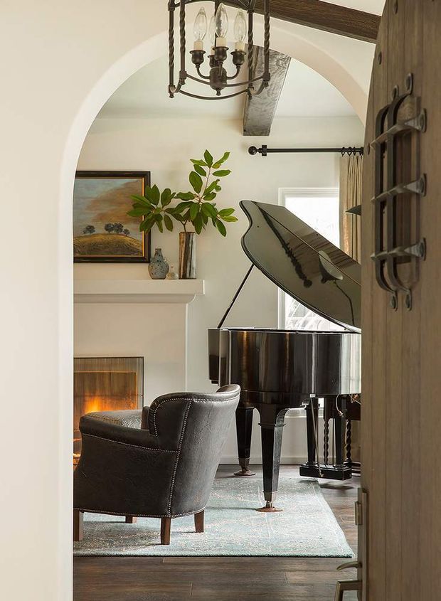 A refined traditional living room viewed through an arched doorway, featuring a glossy black grand piano, leather armchair, fireplace, pale area rug, artwork, greenery, and an ornate hanging lantern. White plaster walls, timber flooring, exposed ceiling beam, and a heavy timber door with dark metal hardware create a warm, classic interior.