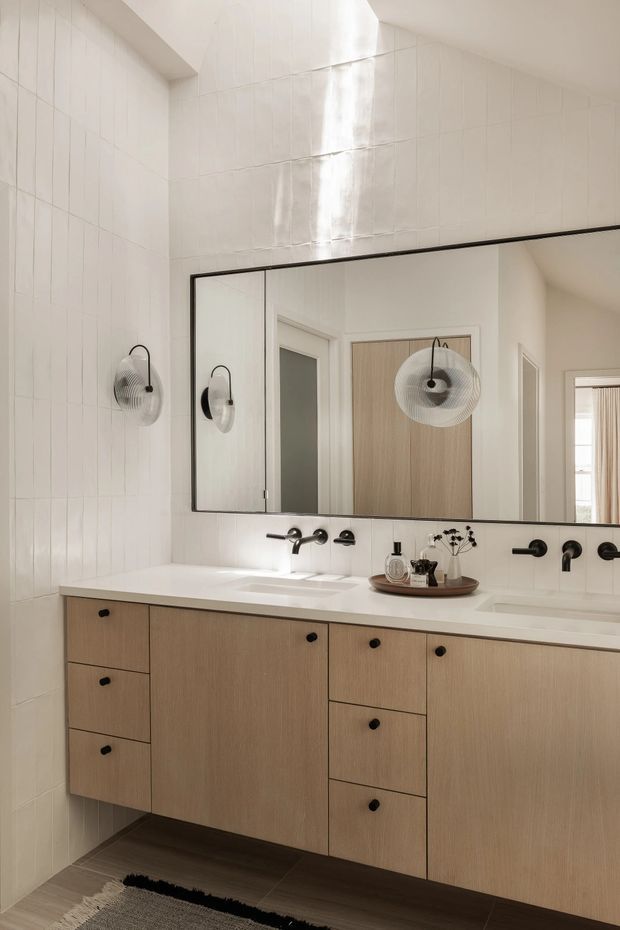 Bright contemporary bathroom vanity with a long double basin, wall-to-wall rectangular mirror, warm light-wood cabinetry, white benchtop and white vertical wall tiles. Matte black tapware, hardware and sculptural wall lights provide contrast, while a dark floor and partial rug add warmth.