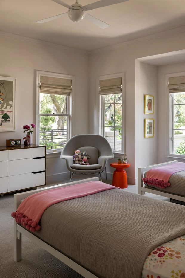 A bright, softly styled bedroom with a white-painted bed, taupe textured bedding, and a pink pom-pom throw. The room has three tall windows with woven Roman blinds, white trim and moulding, a mirrored wall section, a grey upholstered accent chair, a white and wood-front dresser, and a small orange side table. A ceiling fan and colourful framed artwork add character.