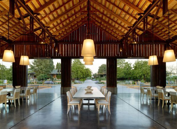 A large open-plan dining and entertaining pavilion with a dramatic exposed timber gable roof, dark timber structural framing and vertical timber wall cladding. Full-height glazing opens views toward a landscaped garden and central water feature. Several timber dining tables and chairs are arranged beneath large warm pendant lights, with a polished dark concrete floor creating a refined indoor-outdoor connection.
