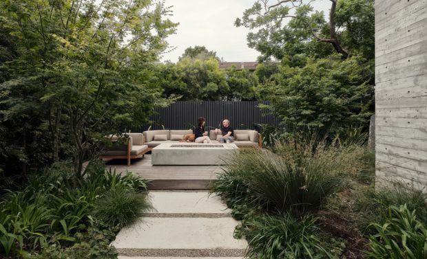 A landscaped outdoor courtyard or terrace viewed from a concrete garden path. A timber deck contains a low outdoor lounge arrangement with a long neutral sofa and central rectangular coffee table, enclosed by dense layered planting and a dark vertical fence. A raw concrete wall forms the right edge of the scene. The setting is calm, private, and contemporary, with naturalistic garden planting and muted earthy tones.