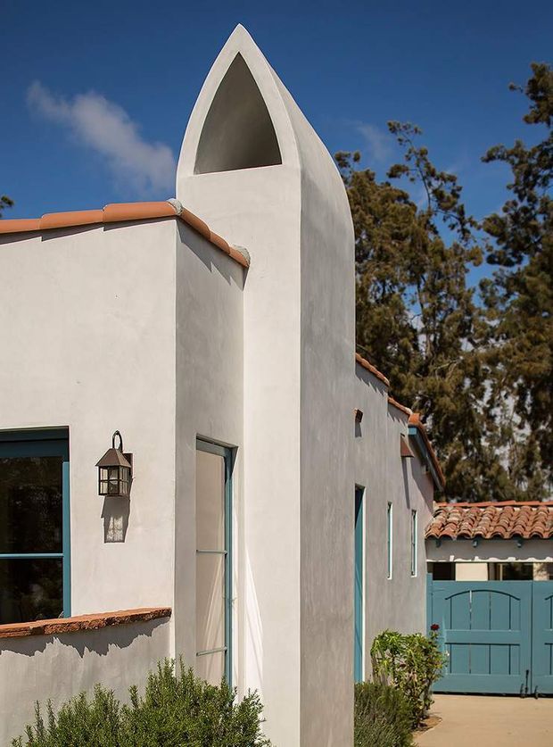 Exterior view of a white stucco building with a tall, pointed architectural tower featuring a recessed triangular opening. The facade has terracotta roof tiles, turquoise-blue window and gate frames, and a traditional lantern sconce. Mediterranean-influenced detailing is visible against a clear blue sky, with shrubs and mature trees around the property.