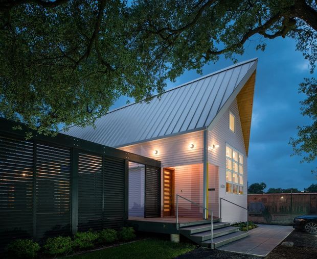 Dusk exterior view of a compact contemporary house with a steep asymmetrical gabled metal roof, light horizontal cladding, warm timber soffit, and a glazed entry facade. A dark slatted privacy screen and covered carport-like structure frame the front, with steps and a paved walkway leading to the entrance. Mature trees surround the property.