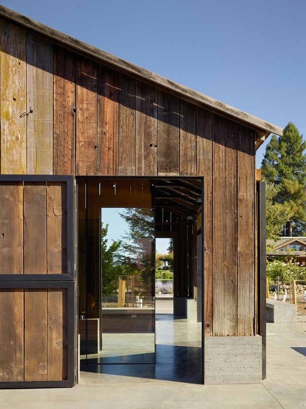 Exterior view of a contemporary barn-like structure clad in weathered vertical timber boards. A large dark-framed glazed entry door opens into a polished concrete-floored interior, with exposed dark timber elements visible inside. The simple sloping roof and rugged timber facade contrast with the crisp glazing and concrete base. Landscaping and another small structure are visible beyond the entrance.