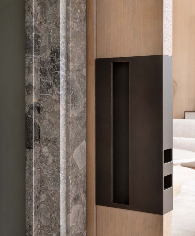 Close-up interior architectural detail showing a dark metal built-in shelving or niche unit integrated into warm vertical timber wall panelling, beside a dramatic grey natural stone feature surface. A light-toned living area is partially visible beyond the opening.