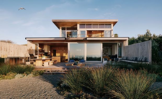 A contemporary two-storey residence viewed from the landscaped garden, featuring broad glazing, warm timber cladding and soffits, a projecting upper level, and an open outdoor kitchen and entertaining terrace. Native grasses and timber privacy fencing frame the home.
