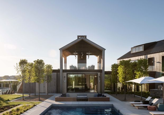 Exterior architectural view of a contemporary two-storey residence centred on a gabled glazed pavilion, opening onto a landscaped courtyard with a rectangular swimming pool, paved terraces, outdoor loungers and umbrellas. A second gabled wing with dark metal roofing and vertical timber cladding is visible to the right. The composition is photographed in bright late-day light.