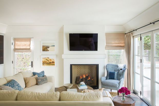 Bright, inviting living room with a white fireplace surround and herringbone-patterned firebox, wall-mounted television, cream sectional sofa, blue upholstered armchair, glass doors, woven Roman blinds, and coastal artwork. The room has abundant natural light and a refined, comfortable residential interior.