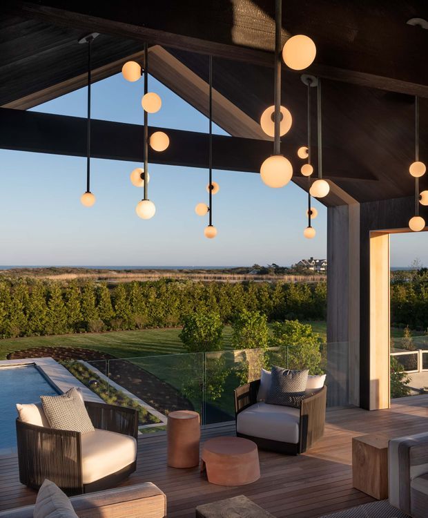 A sophisticated covered outdoor terrace overlooking landscaped grounds, a swimming pool, and a distant coastal horizon. The space features dark timber decking and structural beams, extensive frameless glass balustrading, cushioned lounge chairs, low timber and terracotta-toned tables, and numerous warm spherical pendant lights suspended from a dramatic pitched timber ceiling. A large opening connects the terrace to the interior, creating an indoor-outdoor living area.