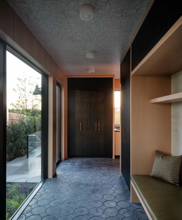 A narrow, carefully detailed interior passage or entry hall with full-height glazing on the left, dark timber doors and cabinetry at the far end, and a built-in upholstered bench with open shelving on the right. The floor is finished in patterned circular tiles, while the ceiling has a repeating embossed geometric pattern. Warm timber and dark wood tones contrast with the cool grey floor and ceiling, creating a refined, dramatic contemporary space.