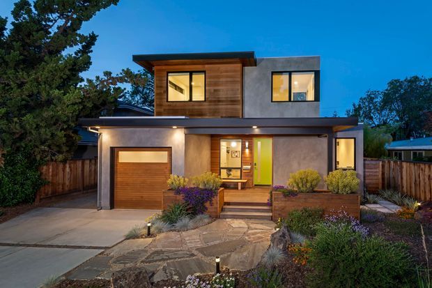 Twilight exterior view of a contemporary two-storey home with a flat roof, projecting upper-level volume, timber-clad facade section, smooth grey rendered walls, large black-framed windows, and a timber garage door. A covered entry porch, warm interior lighting, paved driveway, timber fencing, and landscaped garden beds define the frontage.