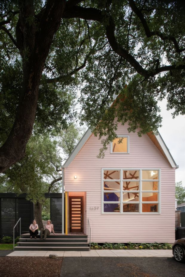 Exterior view of a compact gable-roof house with pale pink horizontal weatherboard cladding, white trim, a modern timber-and-glass front door, and a large multi-pane glazed window. Mature trees frame the building, with stepped entry stairs and a dark slatted boundary fence. Interior spaces and timber structural elements are visible through the front glazing.