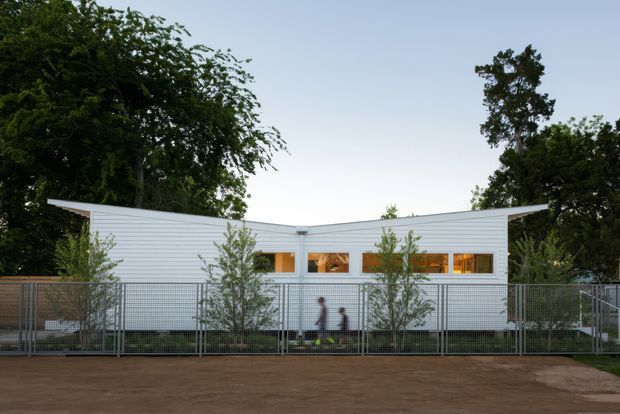 A low, single-storey contemporary building viewed from across a fenced outdoor area. The facade is clad in white horizontal weatherboards, with a shallow asymmetrical roofline and a row of small warm-lit rectangular windows. Mature trees and slender planted trees frame the building, while a metal mesh fence and earthy ground occupy the foreground. Two blurred people provide scale.